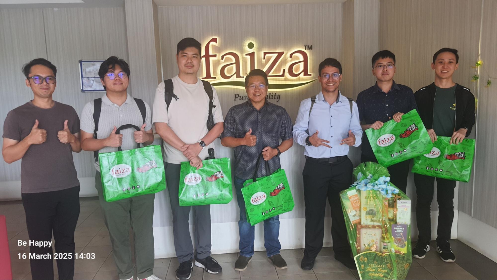 Group photo of staff and visitors posing with Faiza product bags in front of a Faiza company sign.
